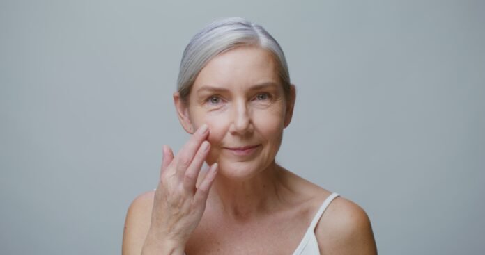 An older model applies moisturizer to her face while smiling at the camera Oily Skin Gets Worse After 35